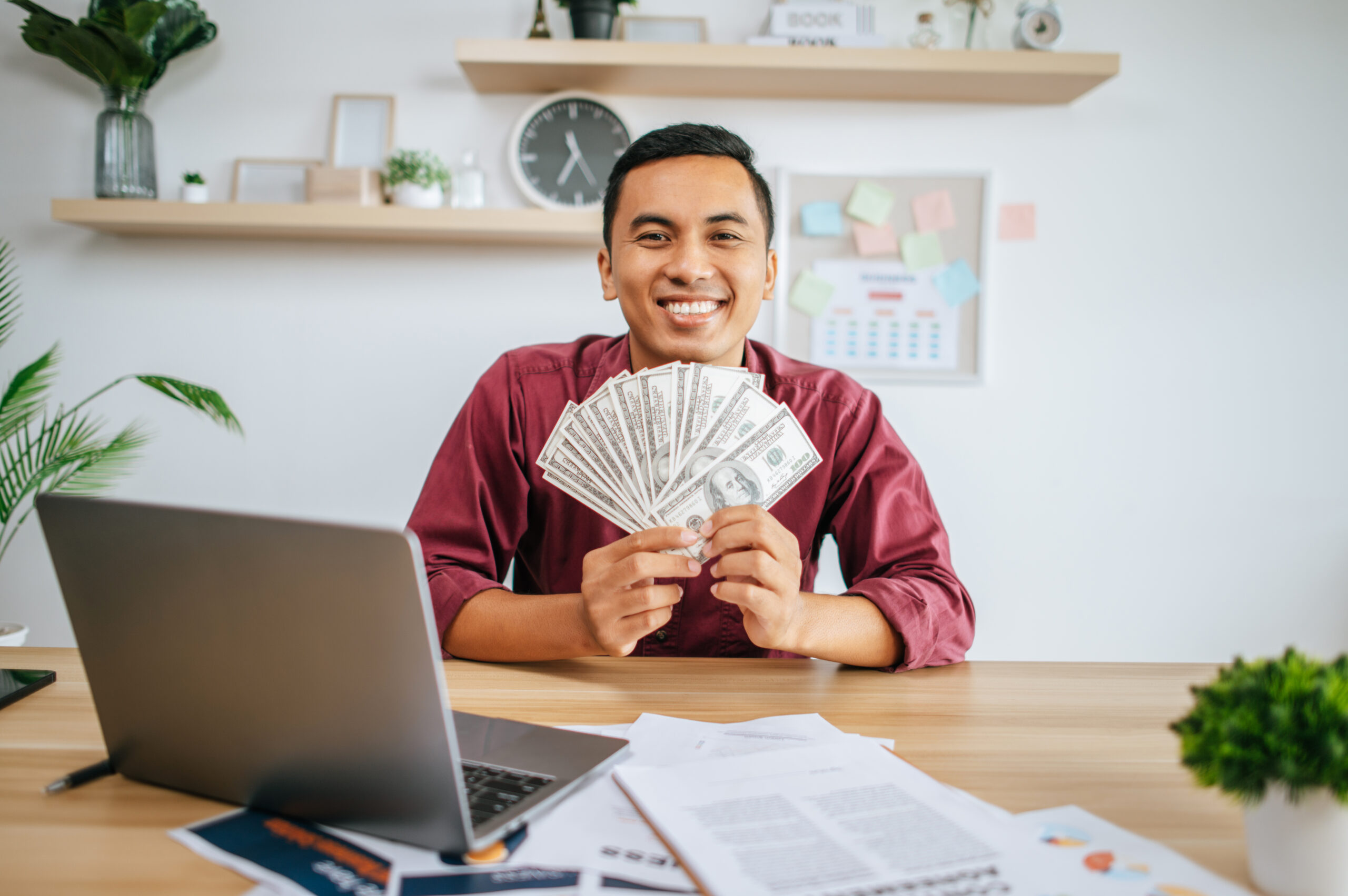a man working in an office and holding money, with a laptop documents on the desk.